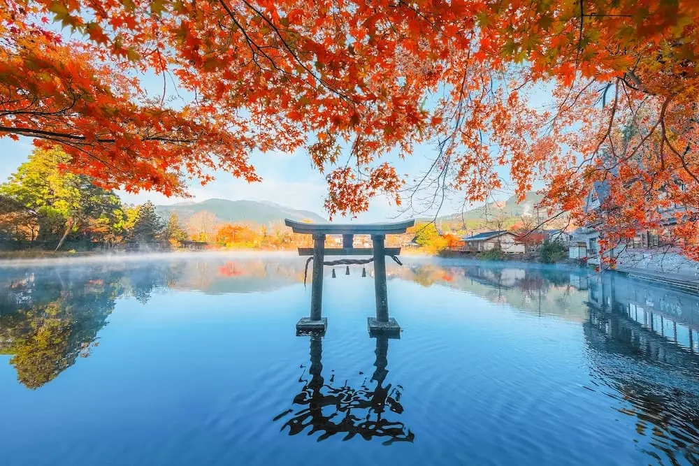 Autumn leaves frame a serene lake with a torii gate at Tenso Shrine in Yufuin, Kyushu, reflecting in the still blue water.