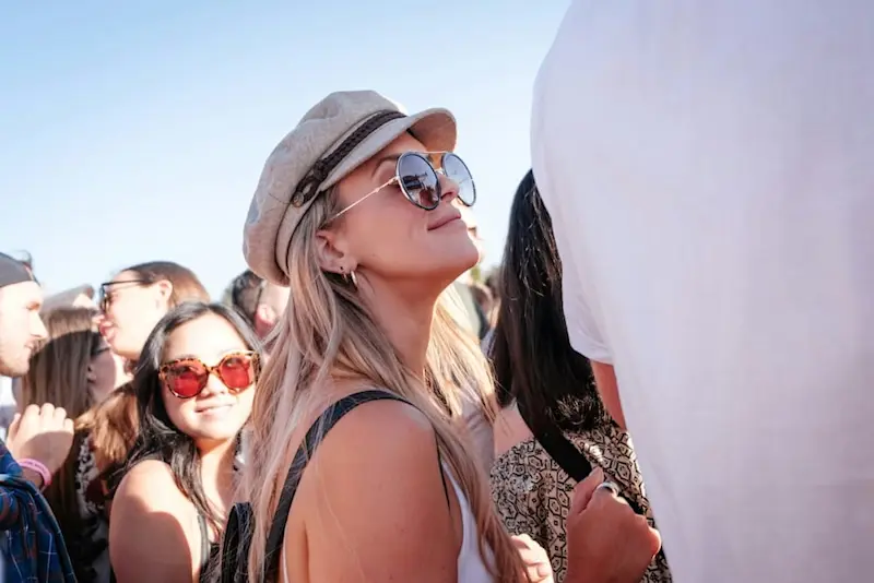 A young woman wearing shades and a cap at St Kilda Beach looking up at the sky, as crowds gather and mingle around her at a party.