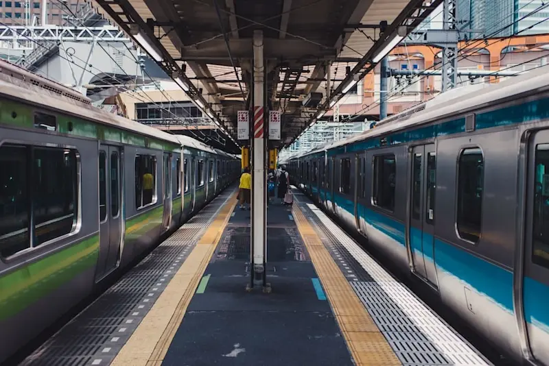 Passengers waiting on a platform in a Japanese train station, with green and blue metallic trains on either side.
