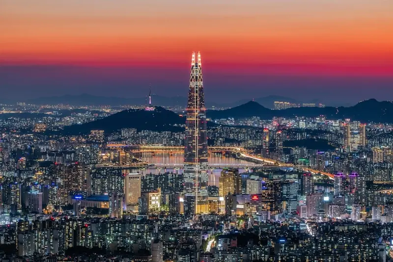 Seoul cityscape at dusk, with towering skyscrapers lit up and the orange glow of the setting sun. Lotte Tower stands as the focal point, with N Seoul Tower visible in the distance on Namsan Mountain.