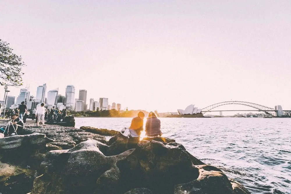 A couple sitting on a rocky coastline, gazing at the distant Sydney skyline with the Harbour Bridge and Opera House, as the warm glow of the setting sun shines between them.