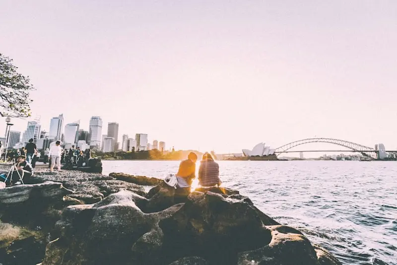 A couple sitting on a rocky coastline, gazing at the distant Sydney skyline with the Harbour Bridge and Opera House, as the warm glow of the setting sun shines between them.