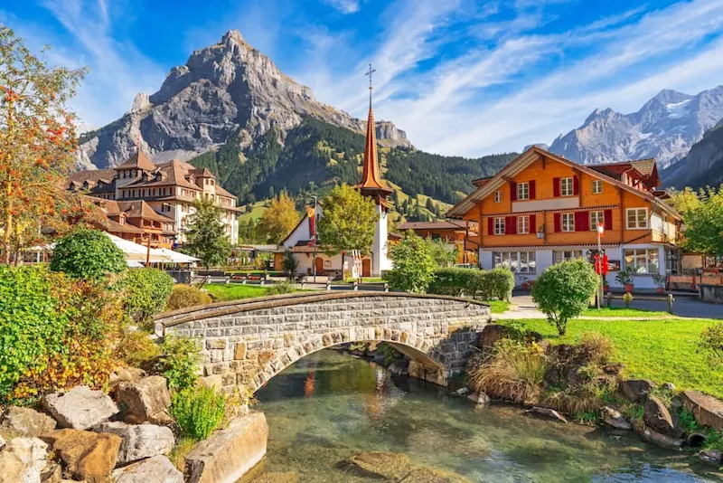 Traditional alpine village of Kandersteg, Switzerland, with wooden chalets, a church spire, stone bridge, and mountain backdrop.