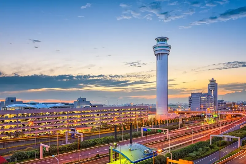Haneda Airport at dusk in Ota, Tokyo, Japan, with the control tower and illuminated terminals against a vibrant sunset sky.