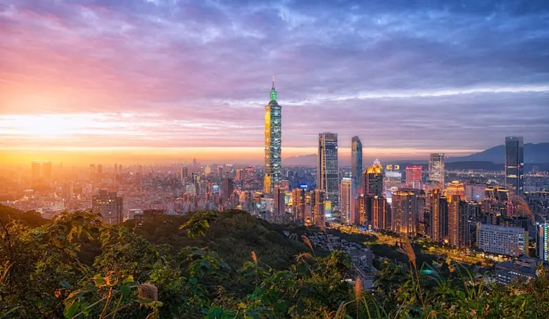 A panoramic view of the Taipei city skyline at sunset from a mountaintop, with the sky painted in orange and purple hues, and the Taipei 101 skyscraper standing tall among other modern skyscrapers.