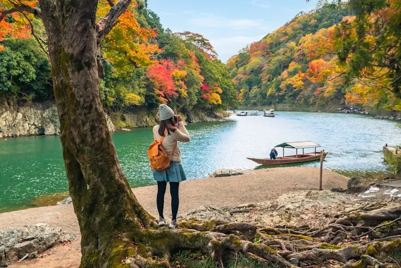 Young woman traveller with an orange bag and a beanie, standing on tree roots and taking a photo of the beautiful autumn landscape in Arashiyama, Japan. Boats float on the river, adding to the scenic view.