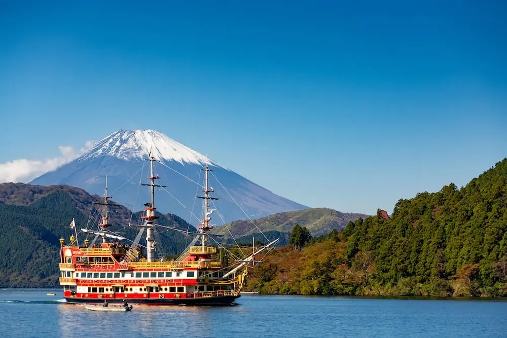A red and yellow Hakone pirate ship cruising across Lake Ashi with Mount Fuji in the background, surrounded by forested mountains under a clear blue sky.