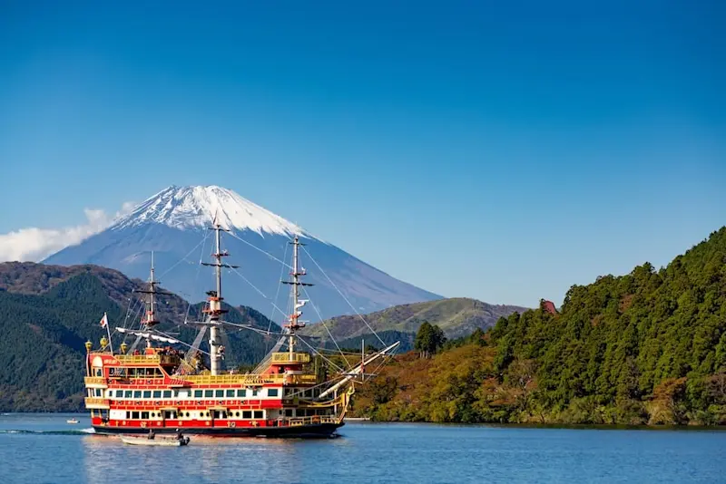 A red and yellow Hakone pirate ship cruising across Lake Ashi with Mount Fuji in the background, surrounded by forested mountains under a clear blue sky.