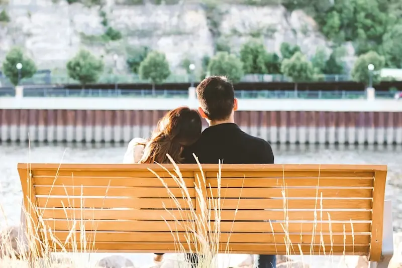 A woman leaning on a man’s shoulder as they sit on a park bench, with flowers swaying in the wind and green trees lined up in front of them.
