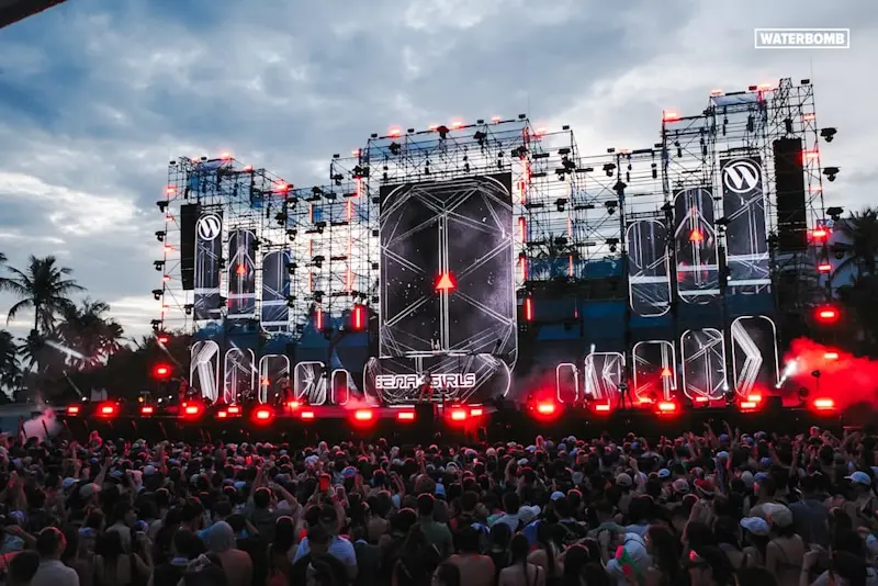 A massive crowd gathers at one of the main stages of Waterbomb Music Festival 2024 in Singapore as the sun sets on a cloudy day, with the stage lights glowing neon red.