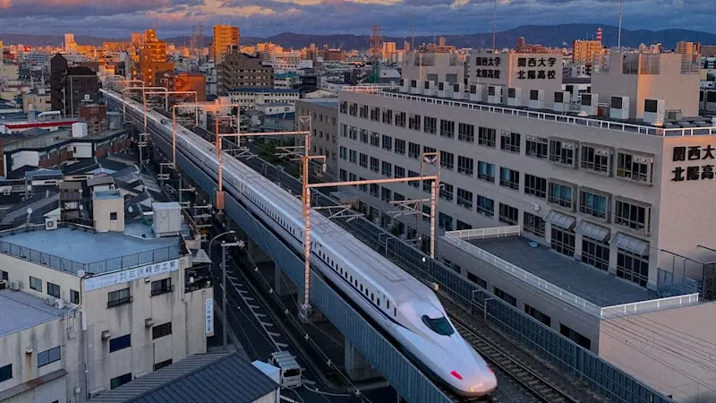 Shinkansen bullet train gliding through urban Osaka at sunset, with city buildings and Kansai University in the background.