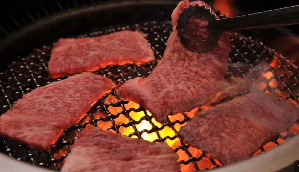 Meat being cooked on a grill, with a pair of tongs holding a piece | Osaka Restaurant