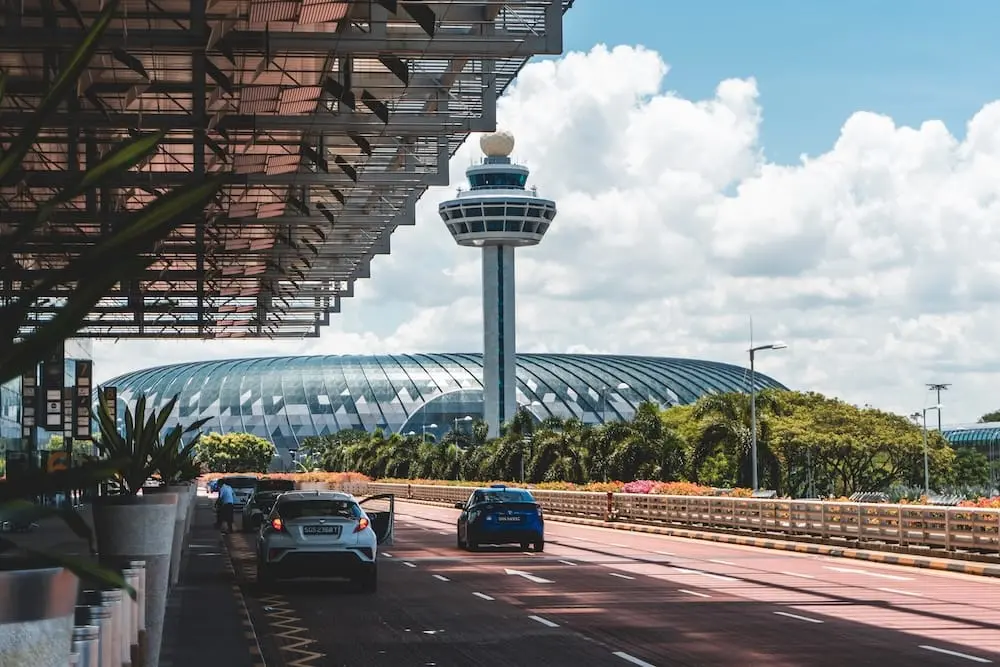 Cars driving past Changi Airport in Singapore with its iconic control tower and Jewel dome under a bright sky with scattered clouds.