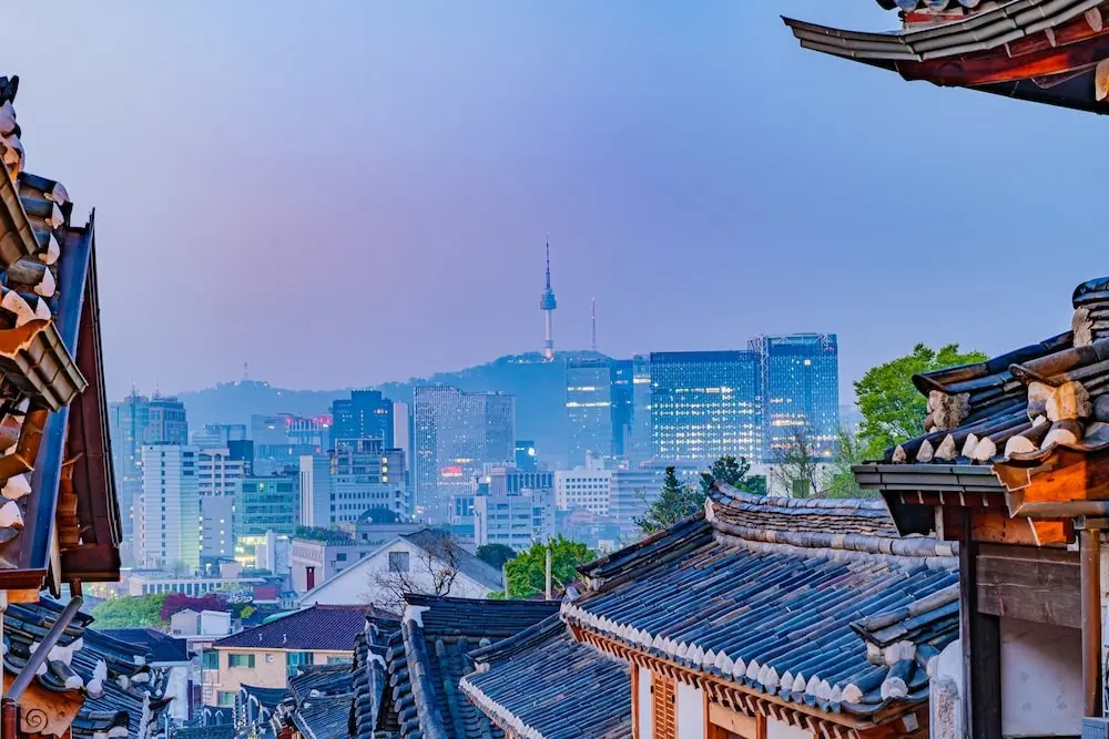 Dusk falls as the Seoul cityscape begins to glitter at night, with the towering N Seoul Tower visible between rows of traditional hanok houses.