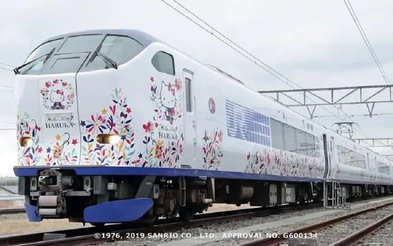Hello Kitty-themed Haruka Airport Express in pink and white with floral designs, travelling on railway tracks in Japan.