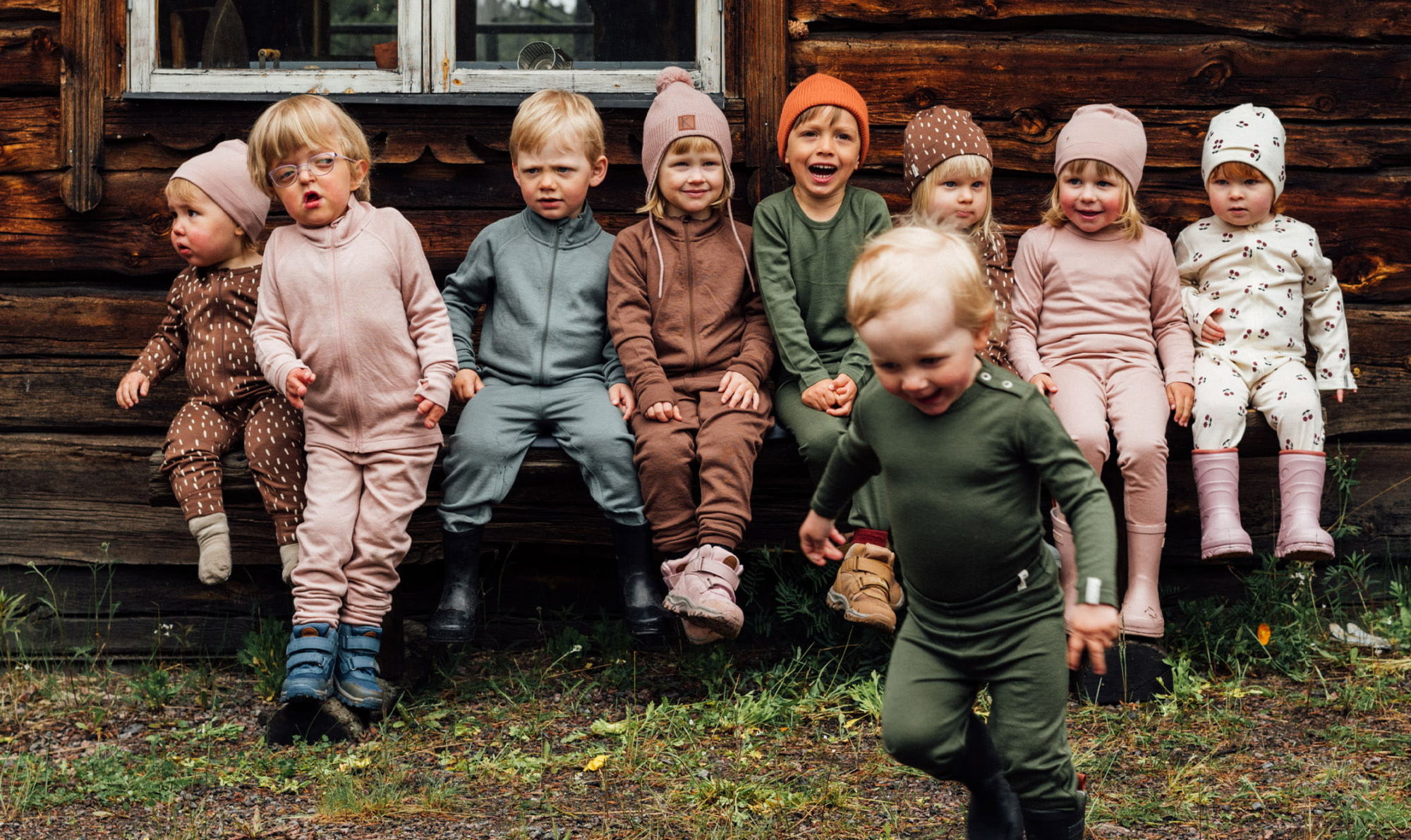 9 kids sitting on a row infront of an old Swedish wooden house in fall clothes - Babyshop