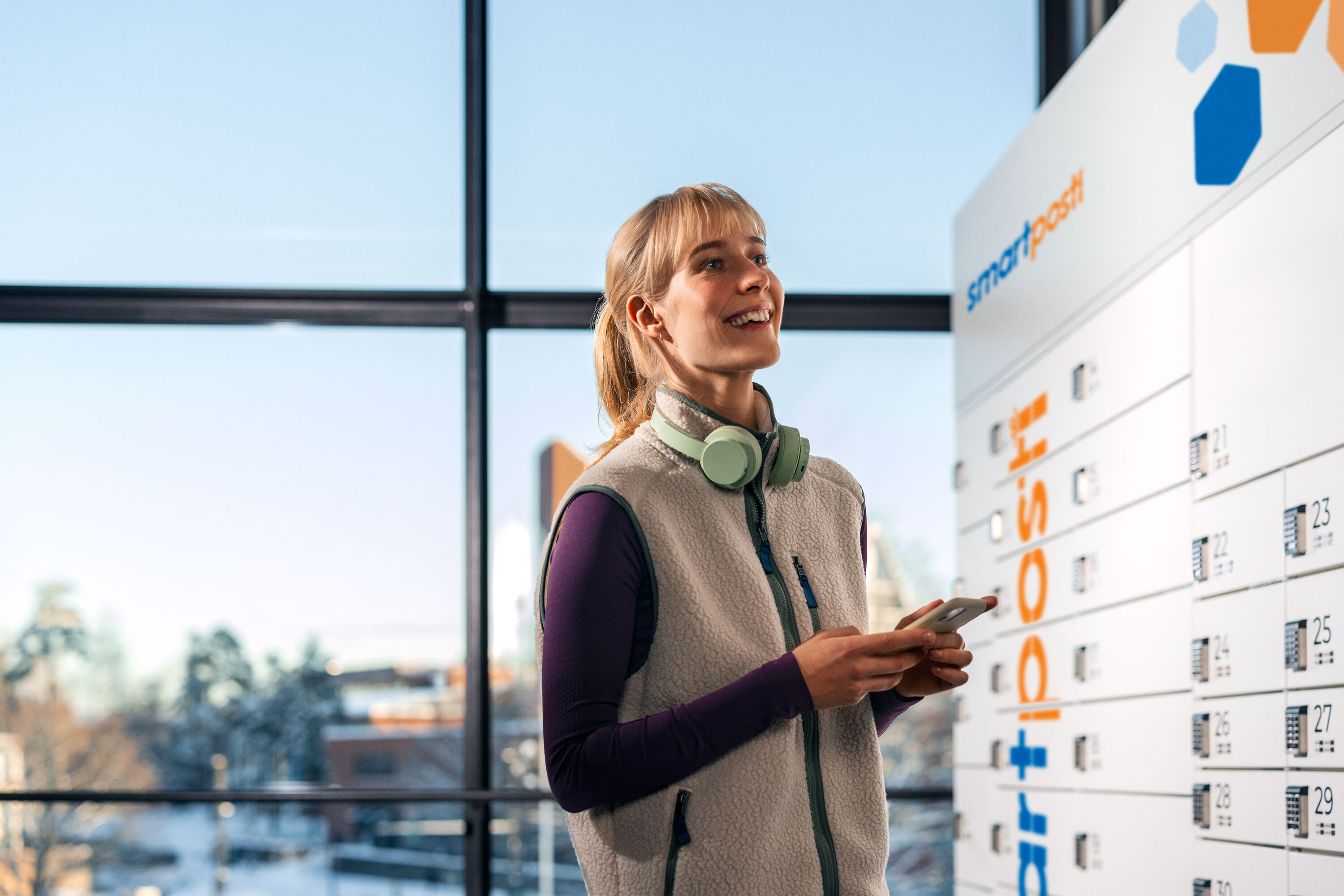 Smiling woman with headphones holding a phone next to a SmartPosti parcel locker machine indoors.