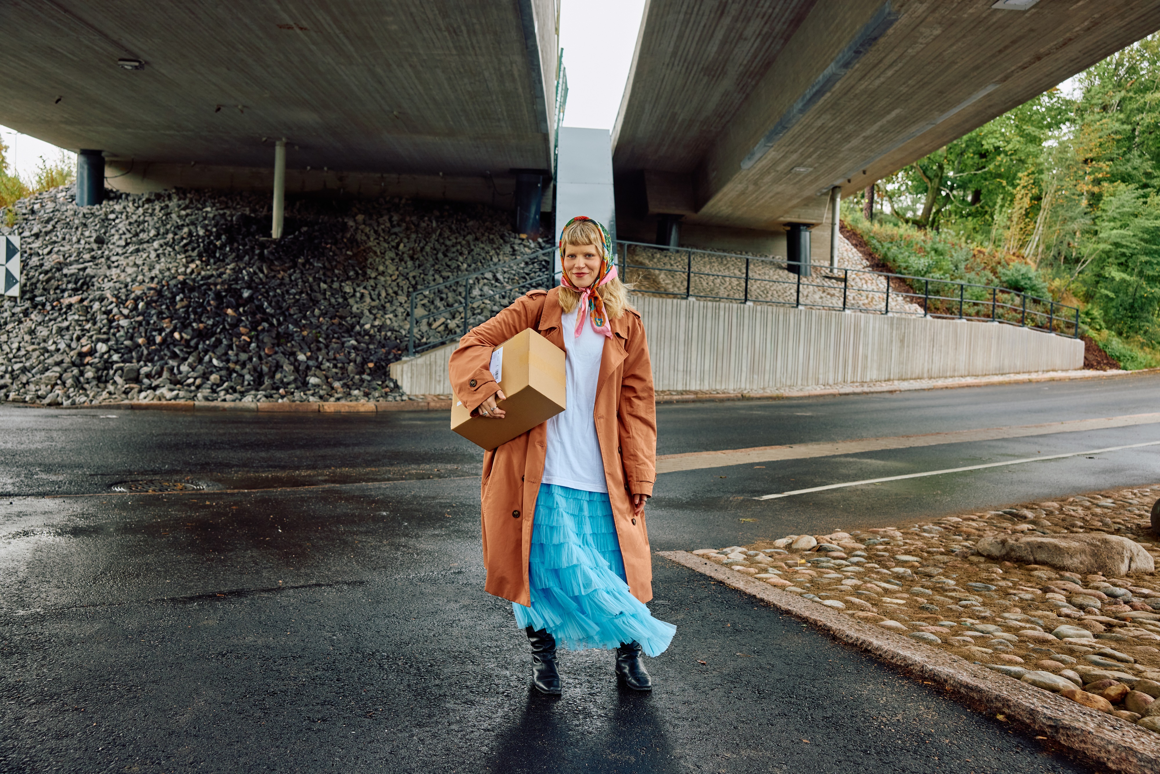 A person stands under a bridge on wet pavement, holding a cardboard box and wearing a brown coat and a blue skirt.
