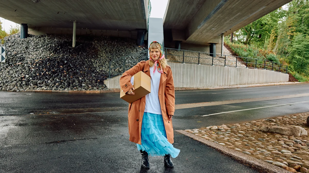 A person stands under a bridge on wet pavement, holding a cardboard box and wearing a brown coat and a blue skirt.