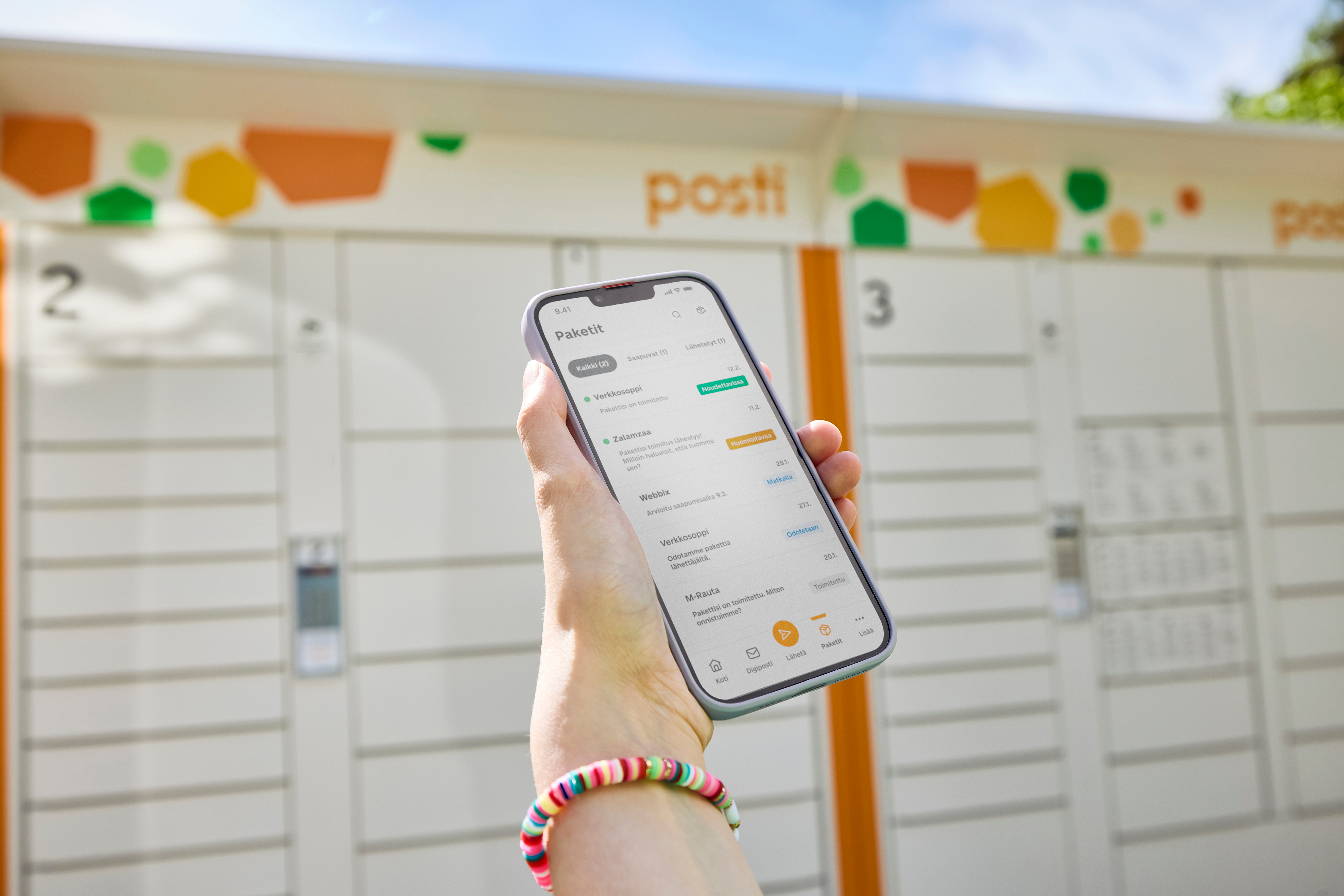 Hand holding a smartphone displaying the Posti app's package tracking screen, in front of a Posti parcel locker station outdoors.