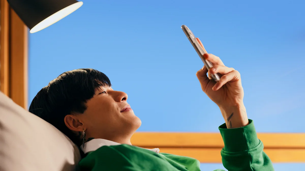 En person ligger i sängen och tittar på sin telefon. Hen har en grön tröja på sig. I bakgrunden syns en blå himmel och fönsterkarmar.
