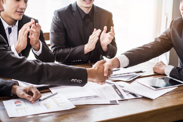 A group of private equity investors sit around a conference room table while two of them shake hands.