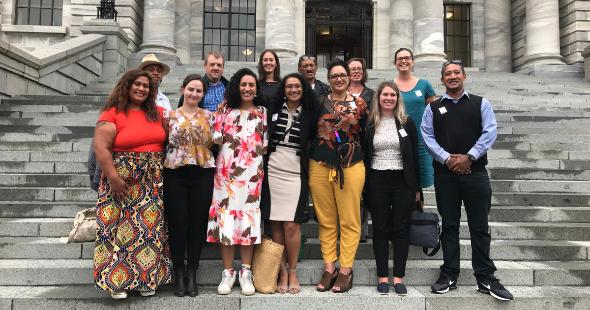 A group of people from different backgrounds stands on the steps of Parliament. They are dressed in bright colours and all have big smiles.