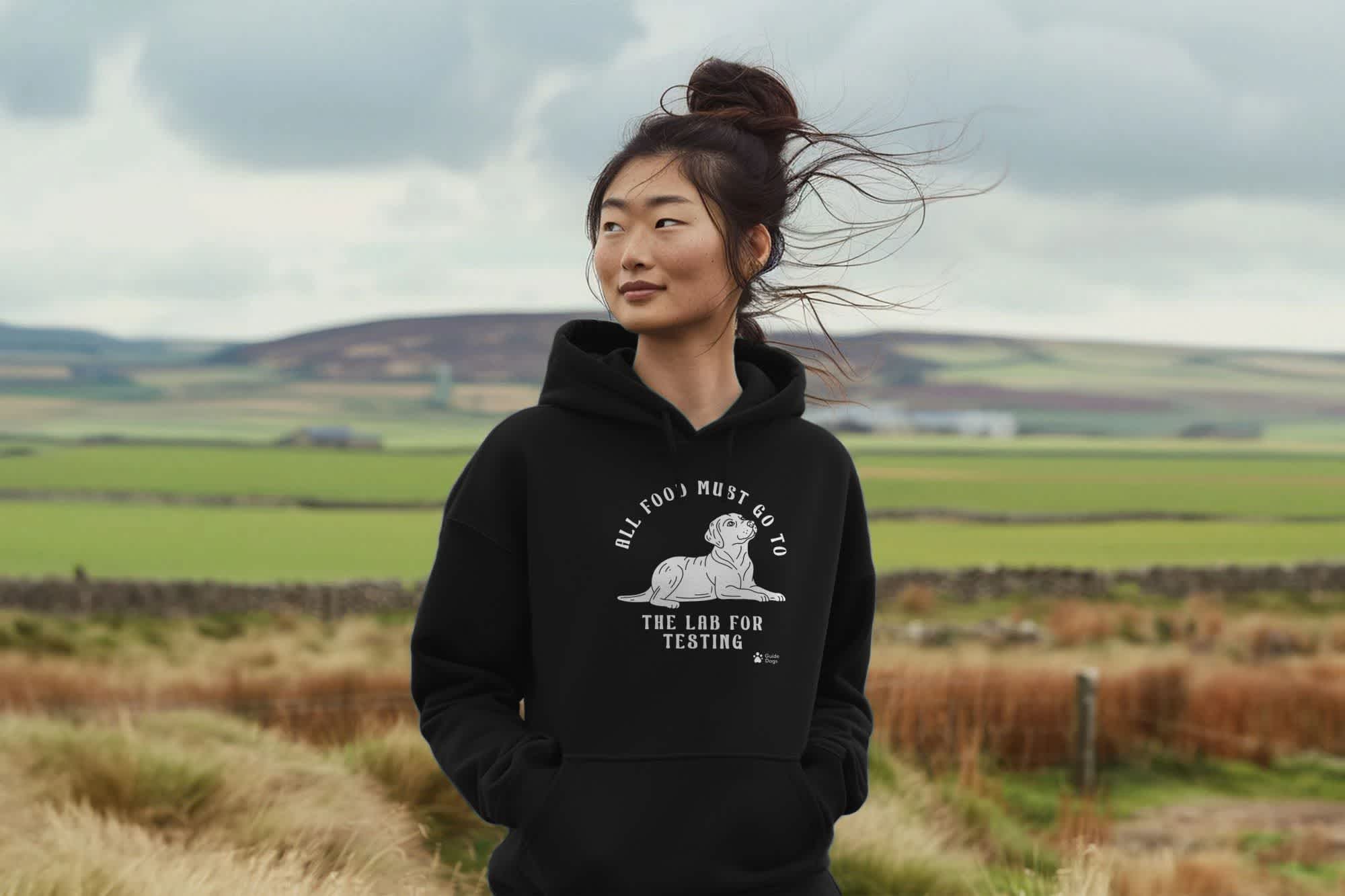 A woman wears a black hoodie with a Teemill Guide Dogs design on it as she looks out into the countryside all around her. 