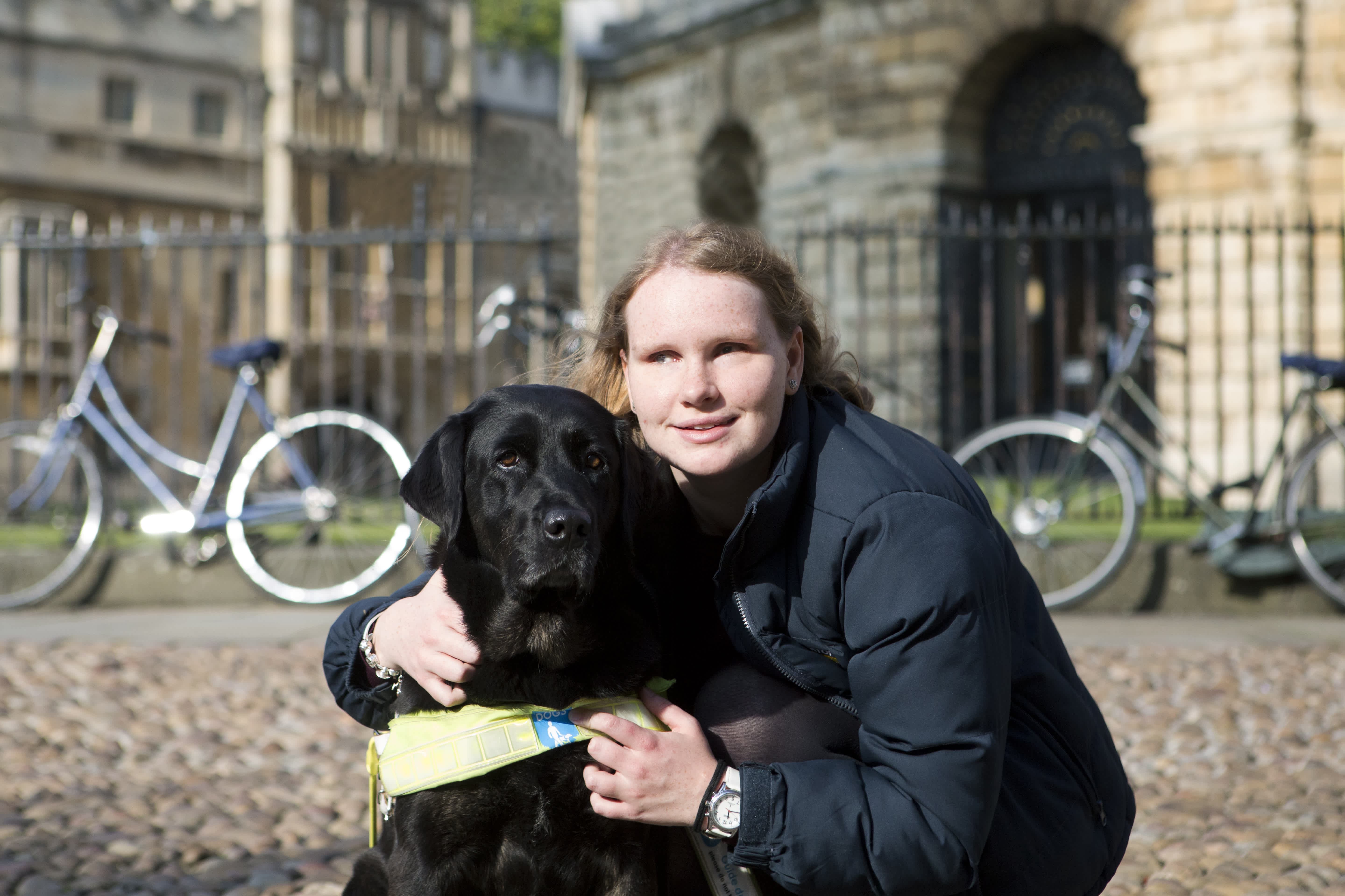 Guide dog owner Ella with black Labrador guide dog Rio crouched down in front of a University of Oxford building. 