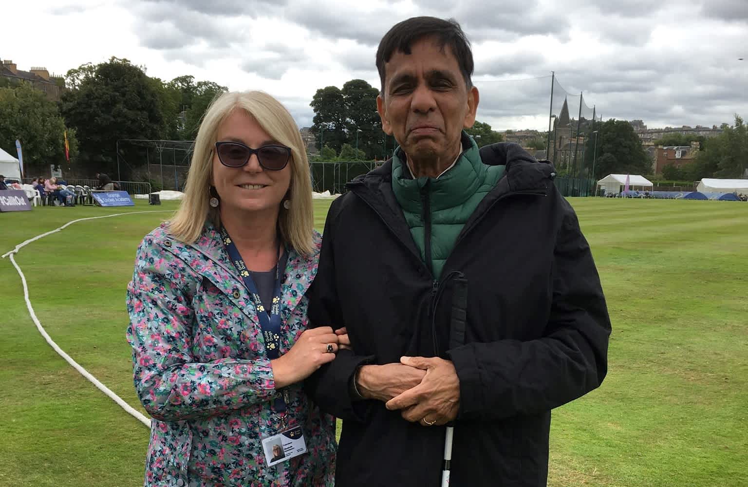 A my sighted guide partnership stand together on the edge of a cricket ground.
