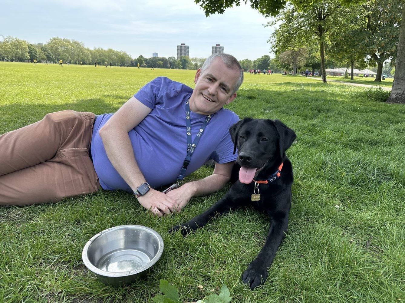 Fosterer Eugene lies on the grass in an empty park as a black Labrador guide dog in training lays beside him with his tongue out and silver water bowl in front of them. 