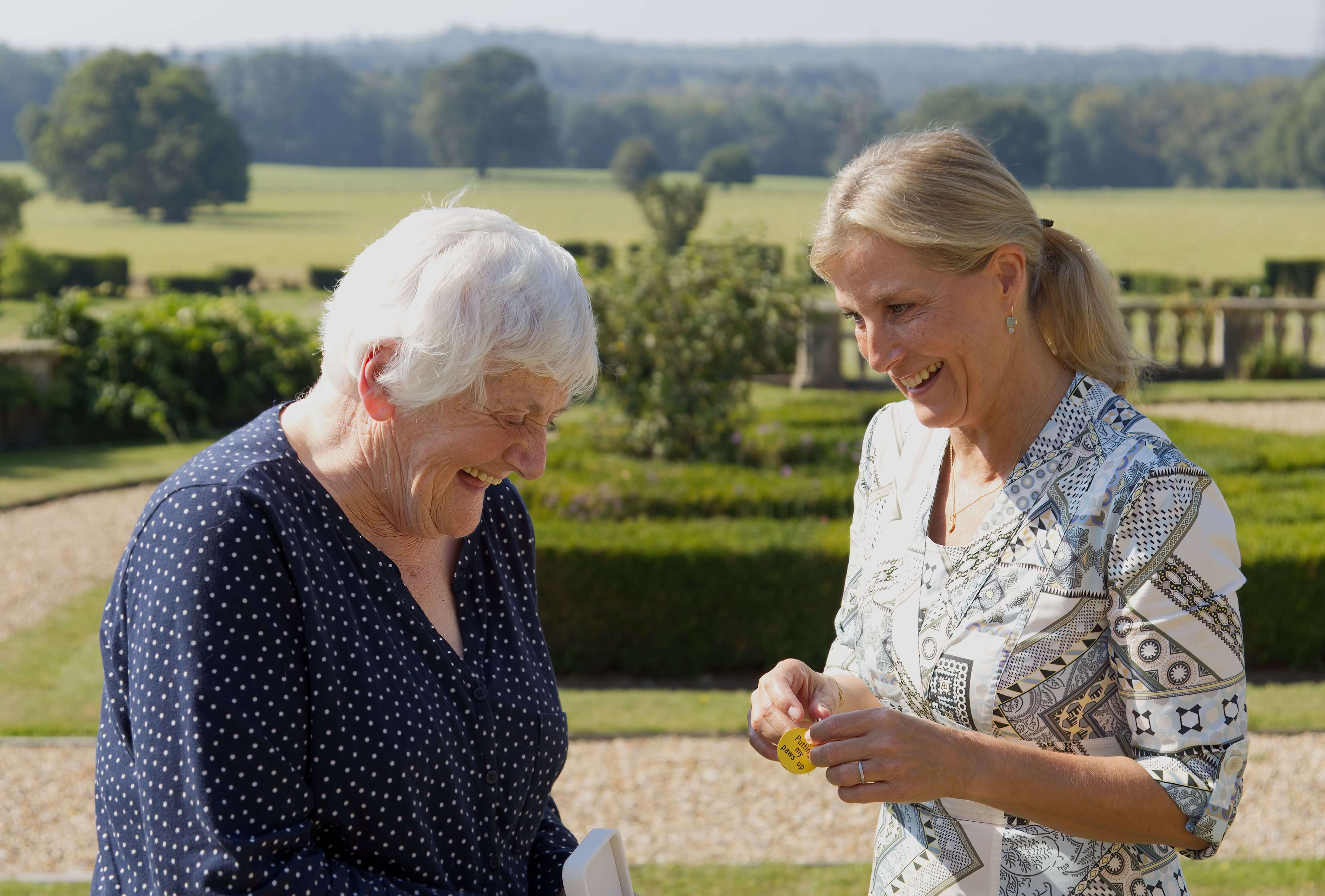 HRH The Duchess of Edinburgh smiles with guide dog owner Mary Pitman. The green landscaped gardens of Bagshot Park are in the background.