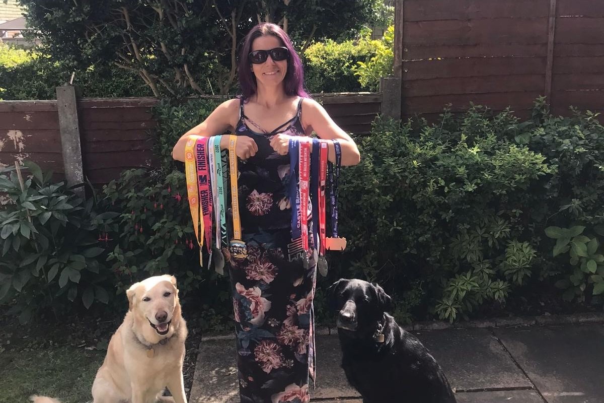 Guide dog owner Laura Whittaker is stood in a sunny garden with her arms draped in running and cycling medals. Sat next to her on one side is her retired guide dog Susan, a Golden Retriever x German Shepherd. On her other side sits her current guide dog Jasmin, a black Golden Retriever x Labrador,