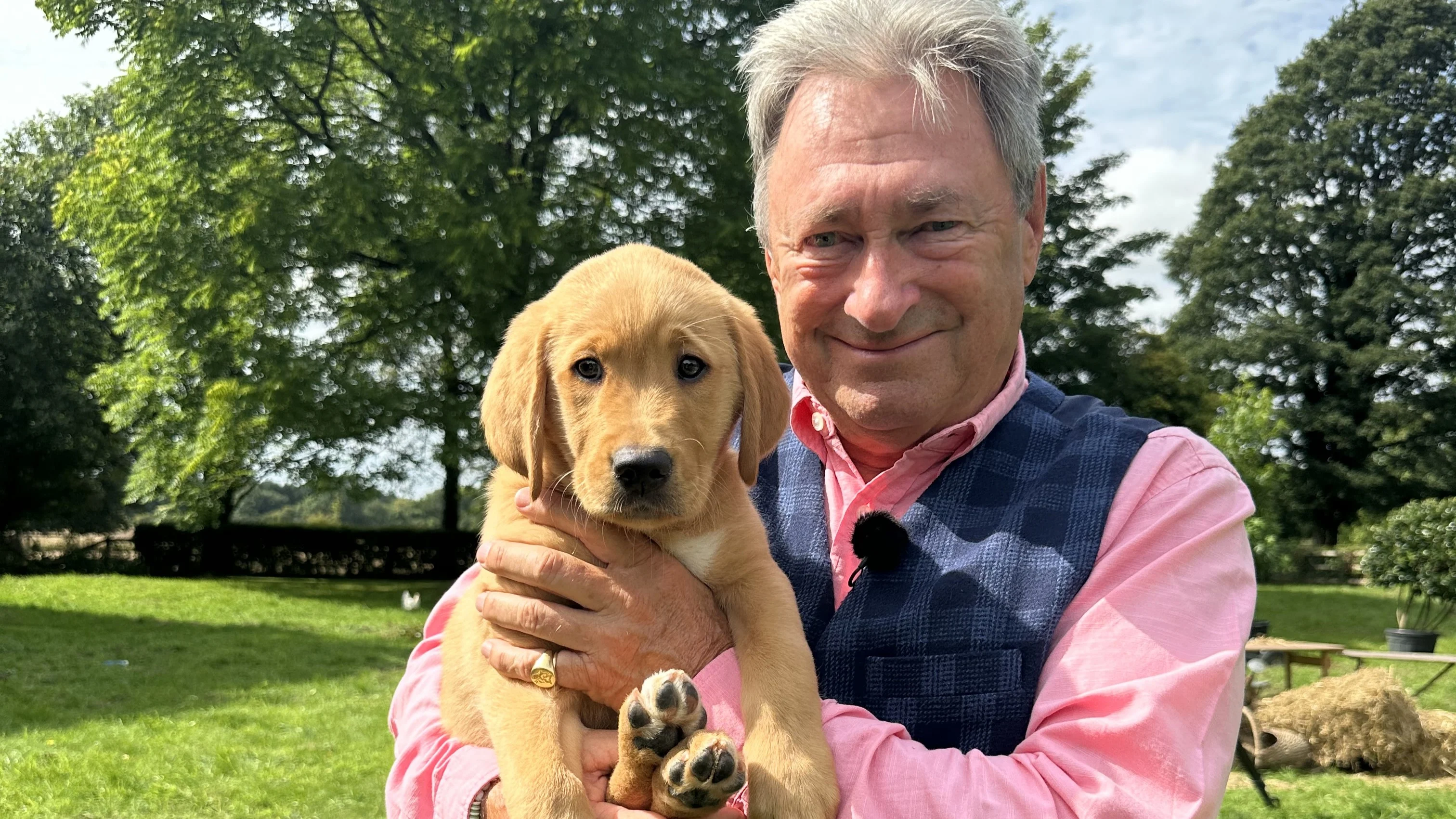 Alan Titchmarsh holds Titch, a golden 9.5-week-old guide dog puppy. Alan wears a bright pink shirt and blue bodywarmer and smiles to the camera.