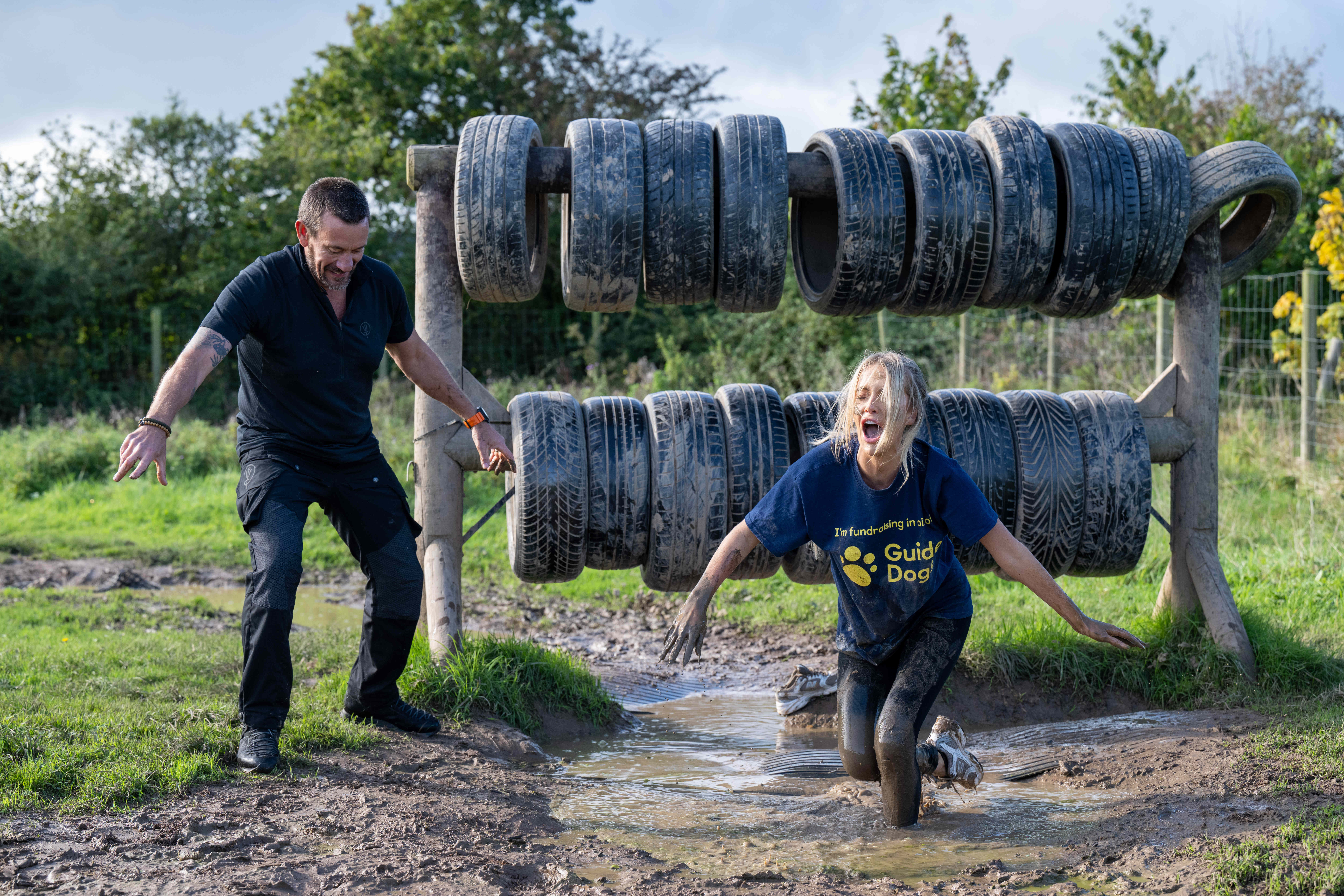 Faye Winter runs through muddy water after climbing over aa wall of tyres as Ollie Ollerton cheers her on. 