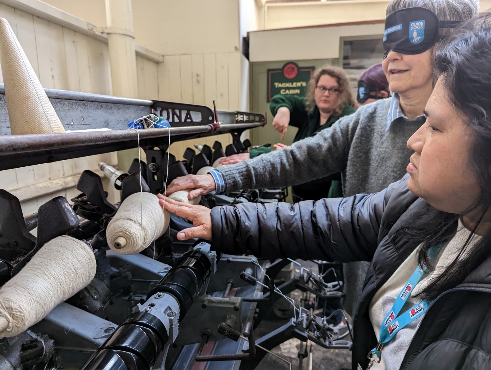 Two staff members from the museum are touching cotton wheels. One person is showing the other where to place their hands, as the second person is blindfolded.