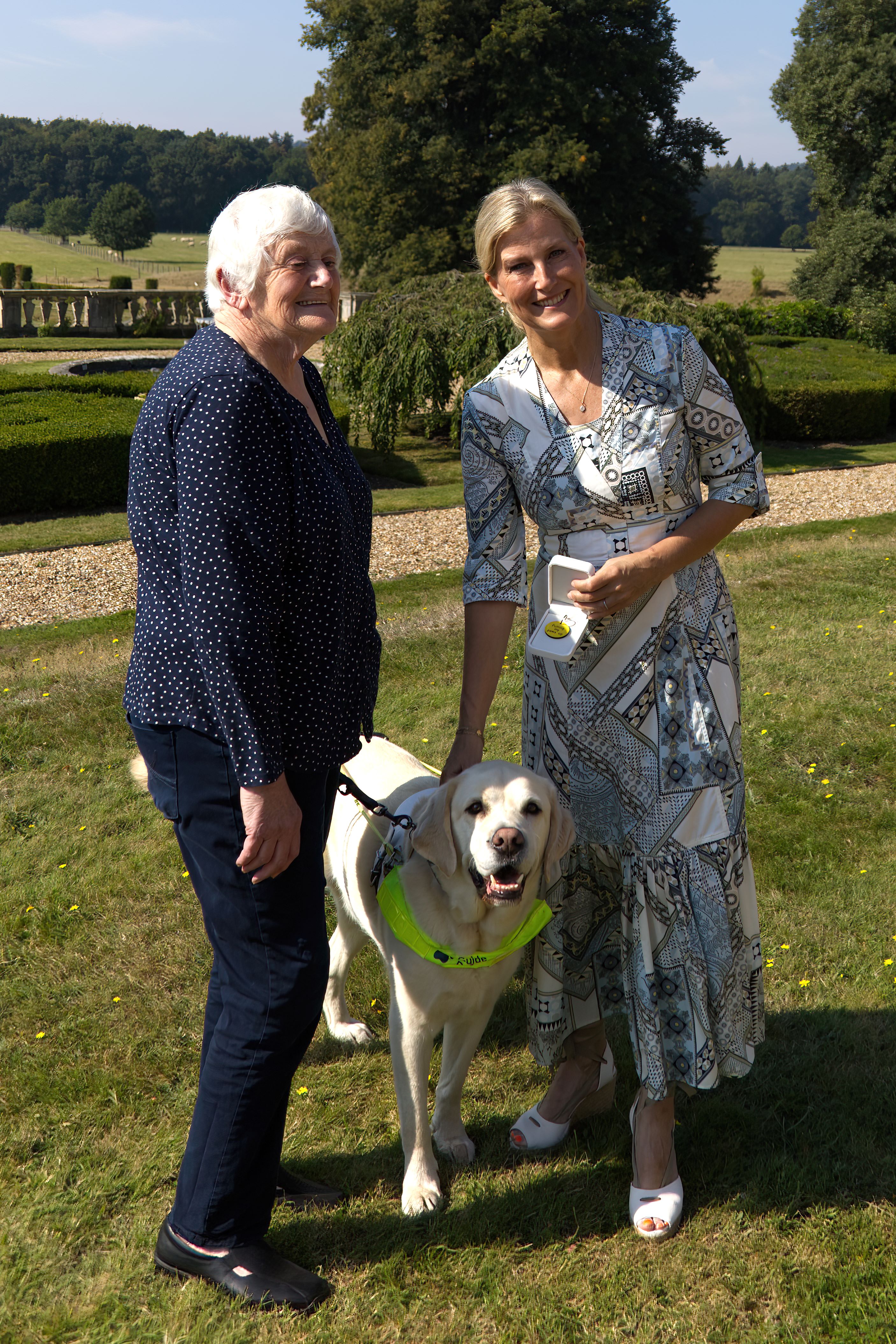HRH The Duchess of Edinburgh stands with Mary Pitman and yellow guide dog Piper in the sunny gardens of Bagshot Park. They all smile towards the camera. 