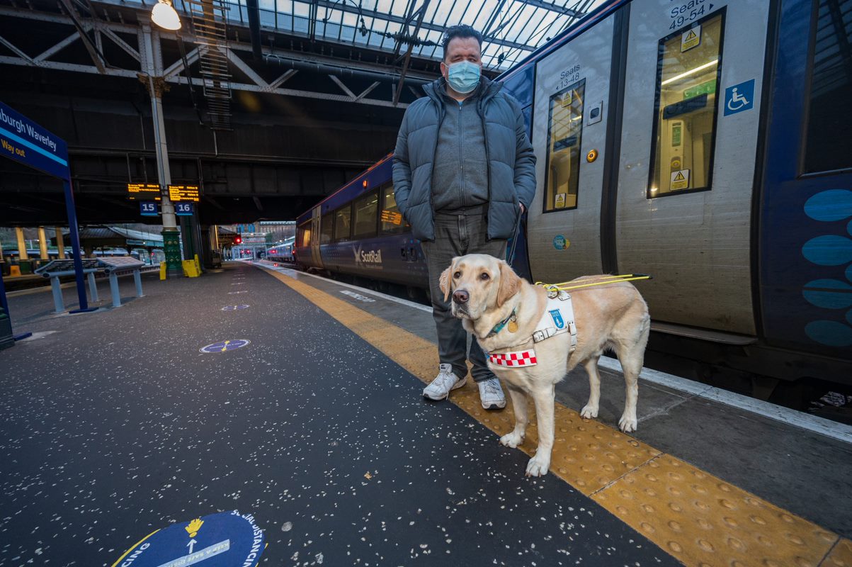 Jon Attenborough on a train station platform with his guide dog Sam.