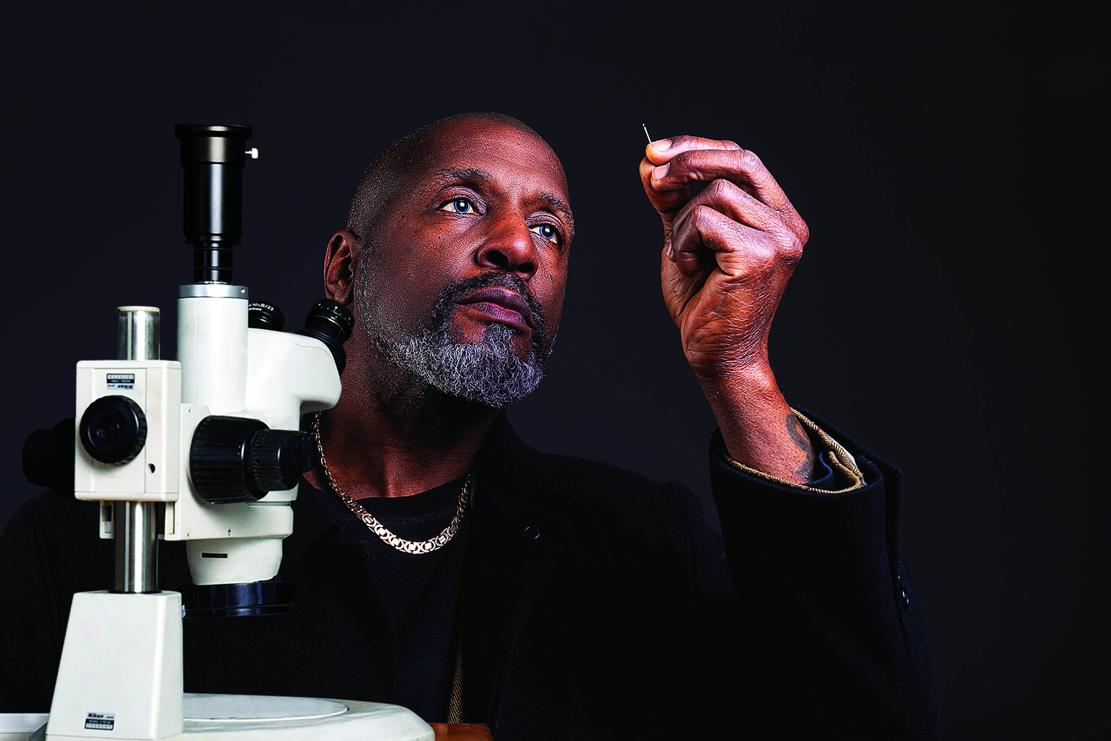 Micro sculptor Willard Wigan holds up a needle to look through the eye at a sculpture as he sits in front of a microscope against a black background. 