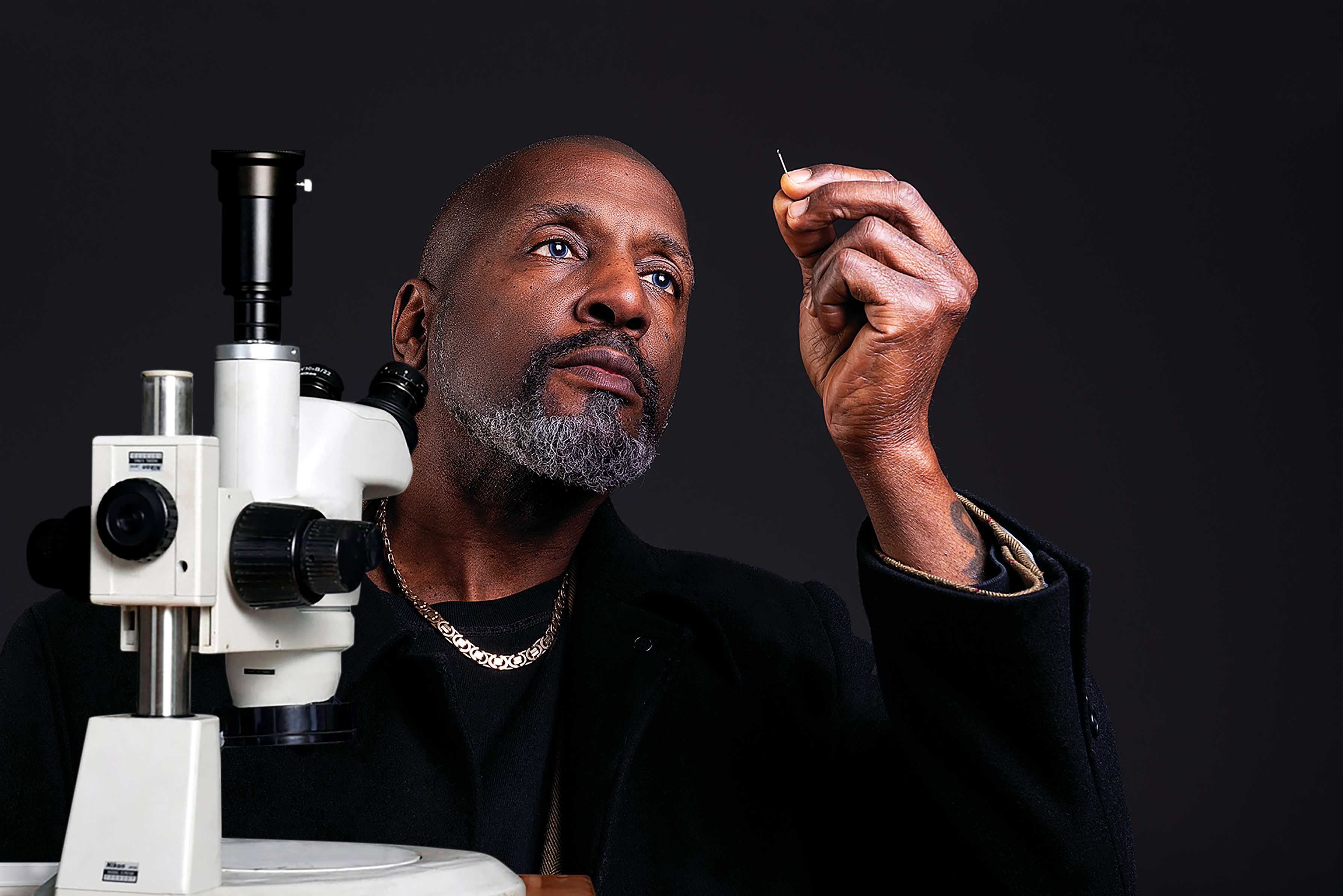 Micro sculptor Willard Wigan holds up a needle to look through the eye at a sculpture as he sits in front of a microscope against a black background. 