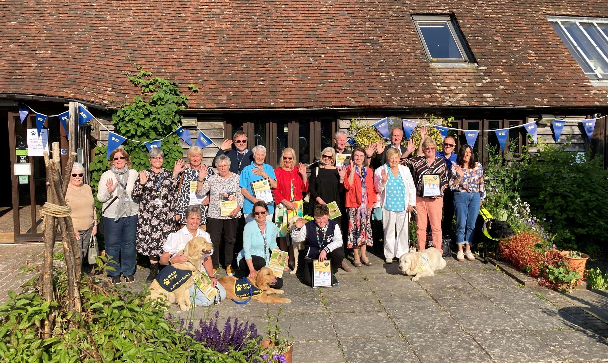 Volunteers, staff and dogs stand outside a barn decorated with bunting and wave at the camera.