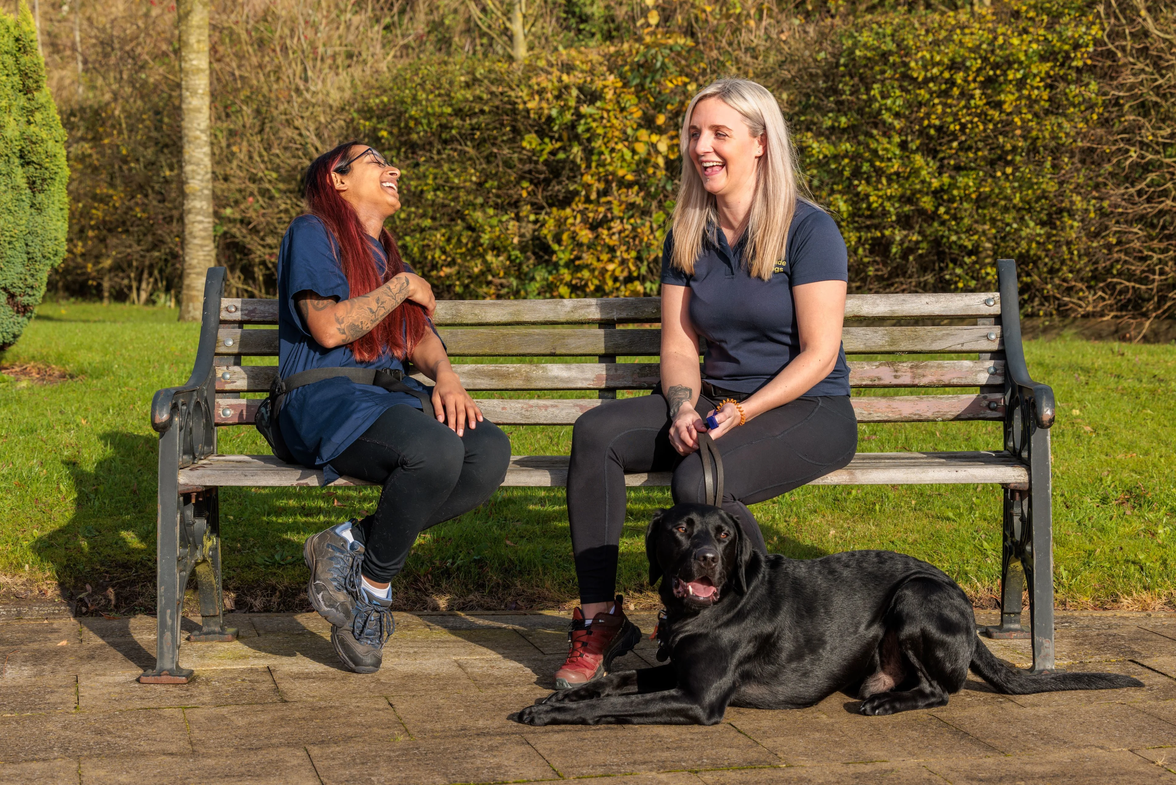 Two women sit on a bench, chatting and laughing. One has a guide dog in training laying at her feet.