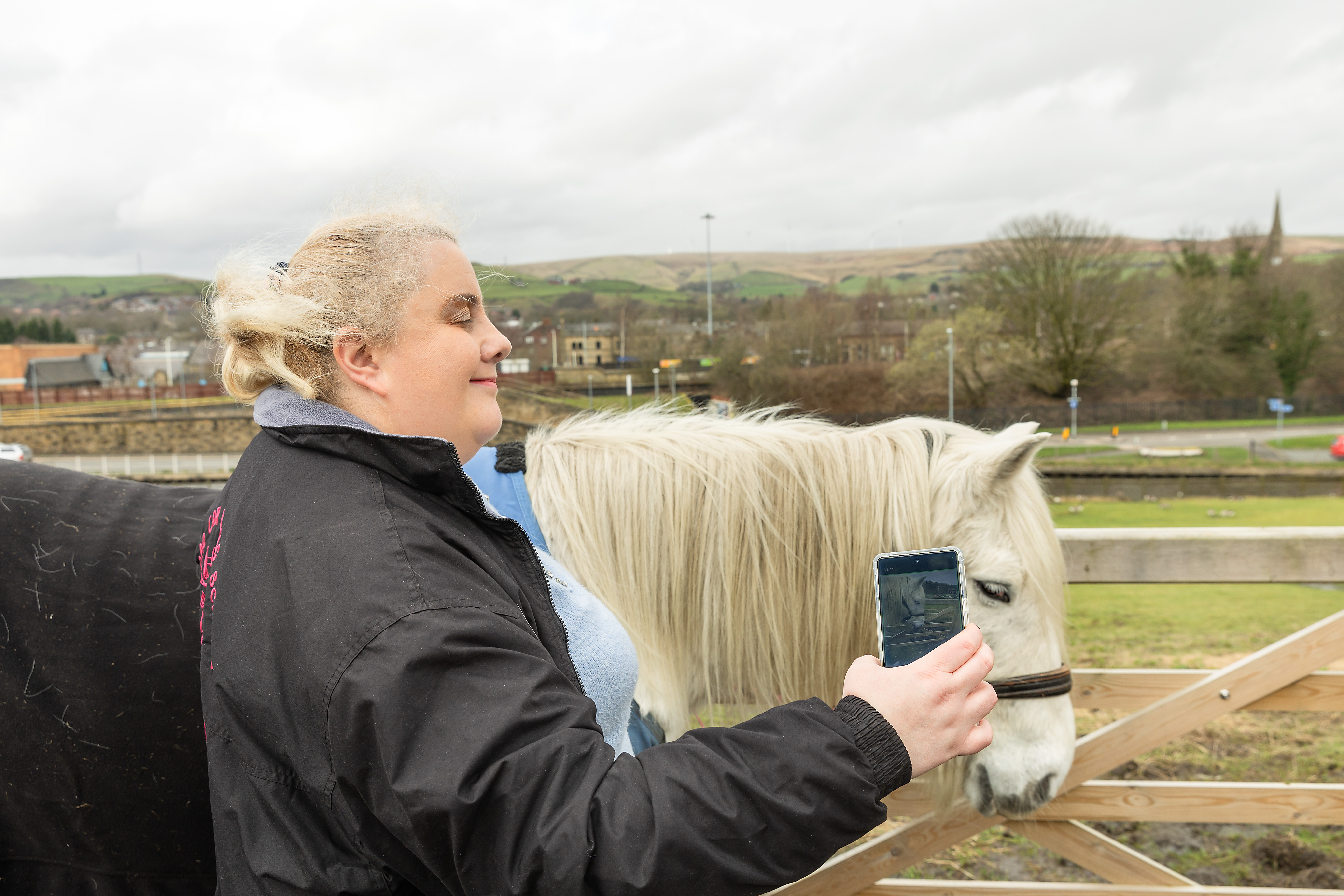 Marie uses her smart phone to take a photo of her horse, stood outside. A field, houses and trees are in the background. 