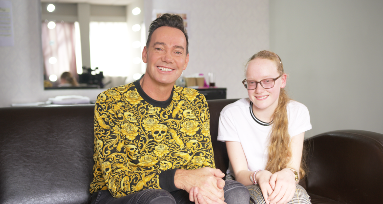 Neve sits on a sofa next to Strictly Come Dancing judge Craig Revel Horwood as they both smile towards the camera. 