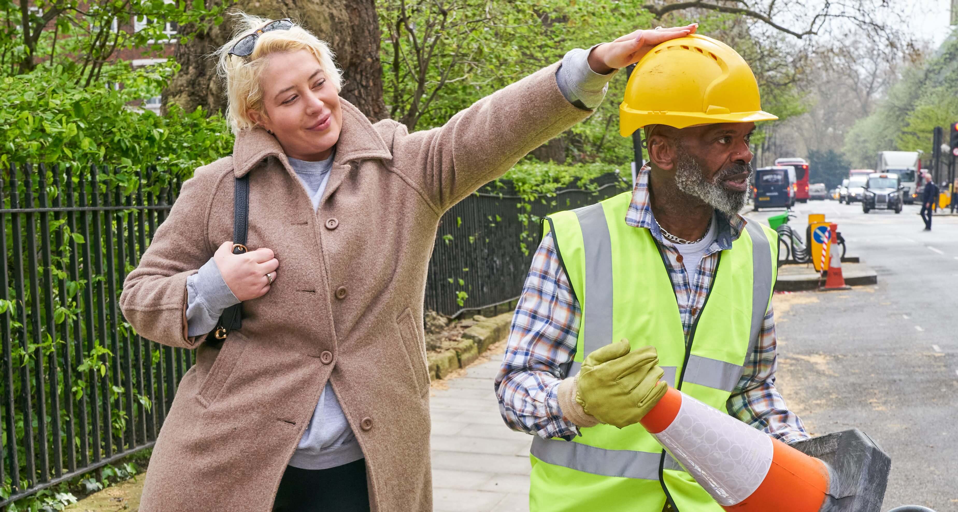 A London street. A woman in a beige coat reaches out and pats the head of construction worker, who is wearing a hard hat and high-vis vest and is laying traffic cones out in the road.