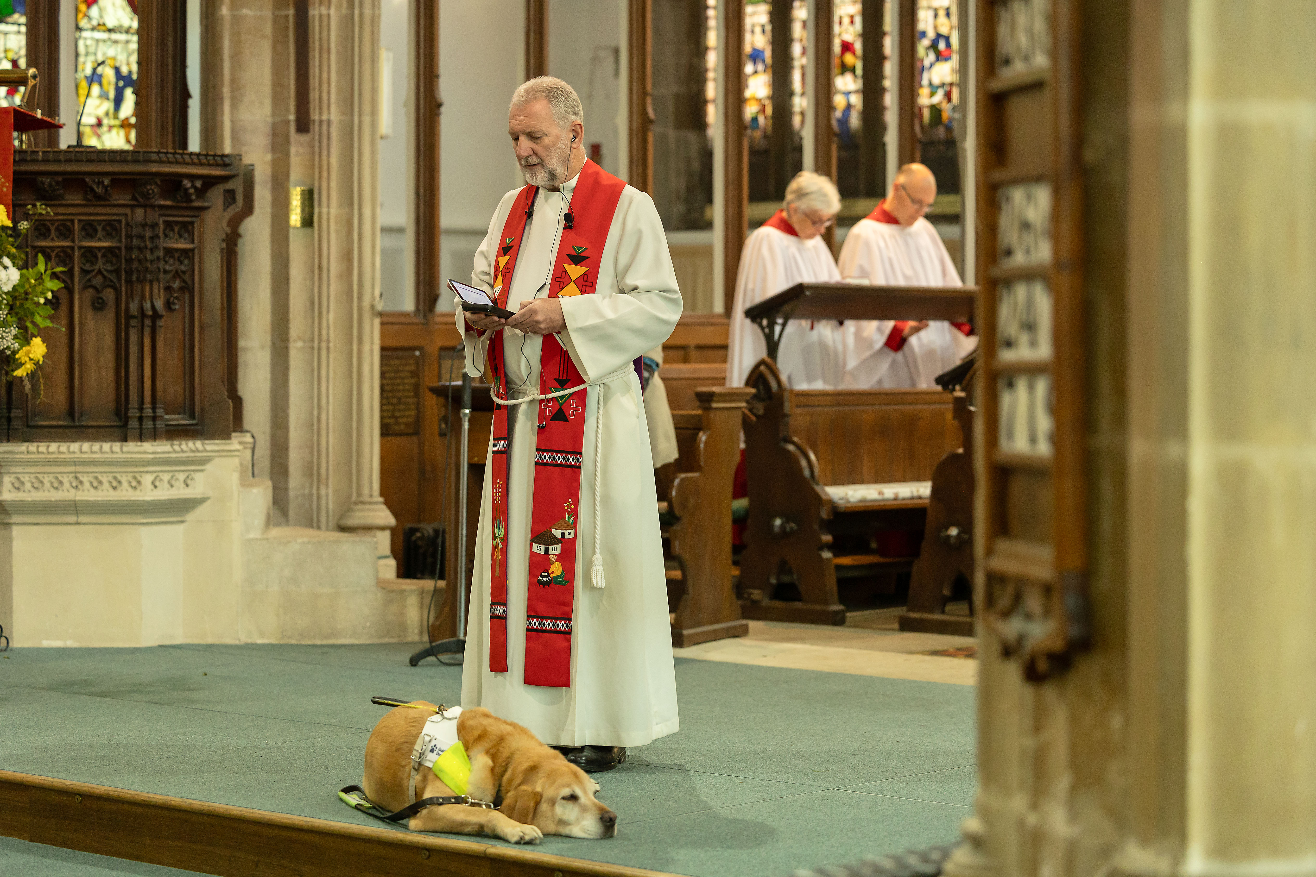 Vicar Simon stands and reads at the altar in church with guide dog Mayne lying at his feet.