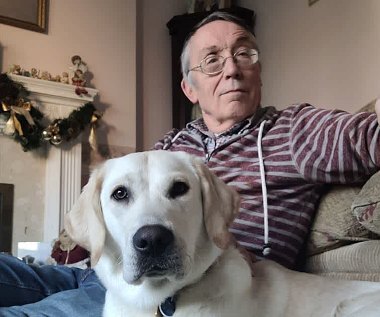 Puppy Raiser Paul sits on the floor with his Labrador puppy lying in front. Paul is taking a photo of the both of them as they are looking at the camera. 