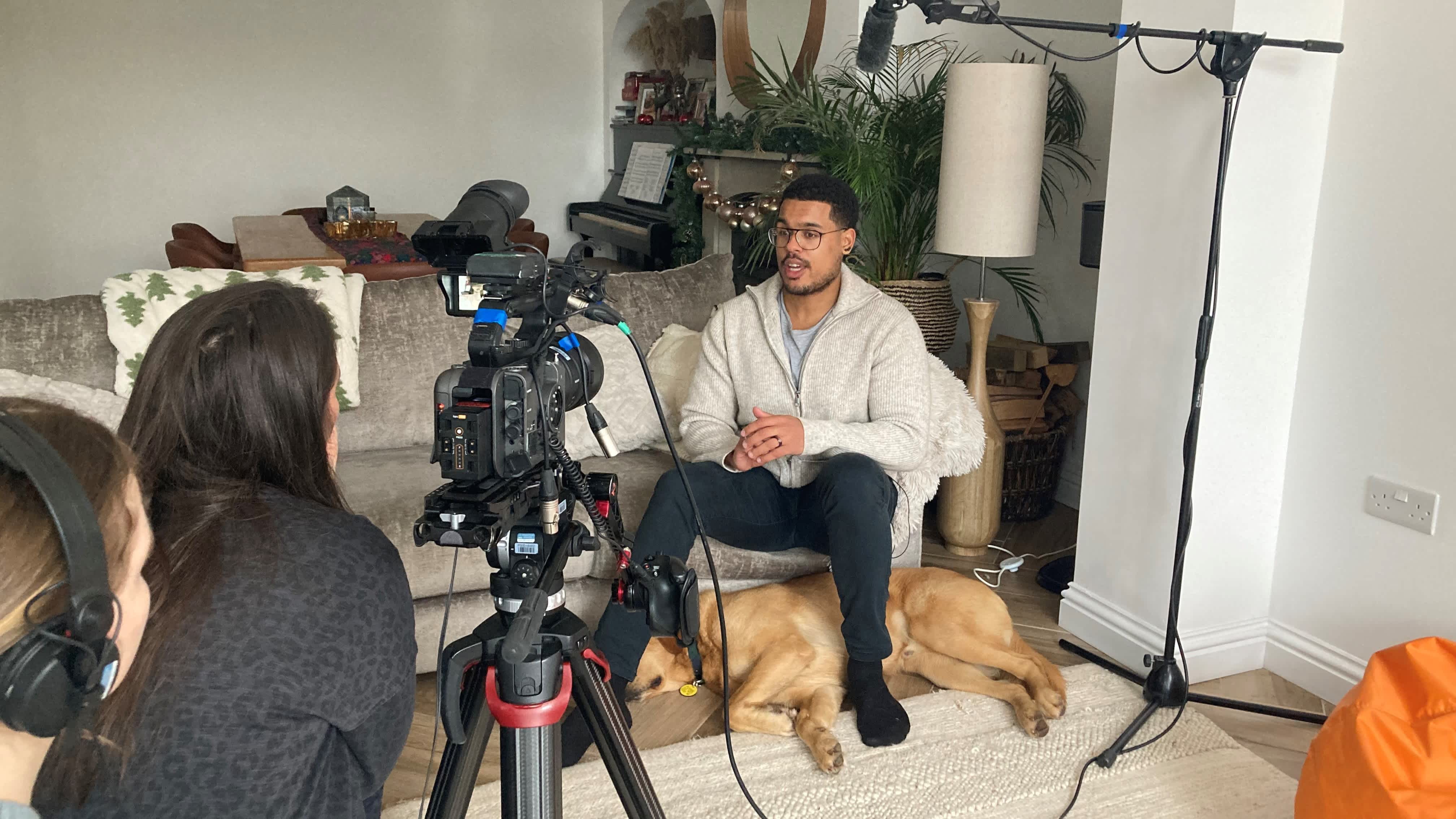 Guide dog owner Devante sits on a grey sofa at home being filmed for BBC Lifeline appeal. His golden guide dog lays at his feet as Devante talks to the camera and two crew members.