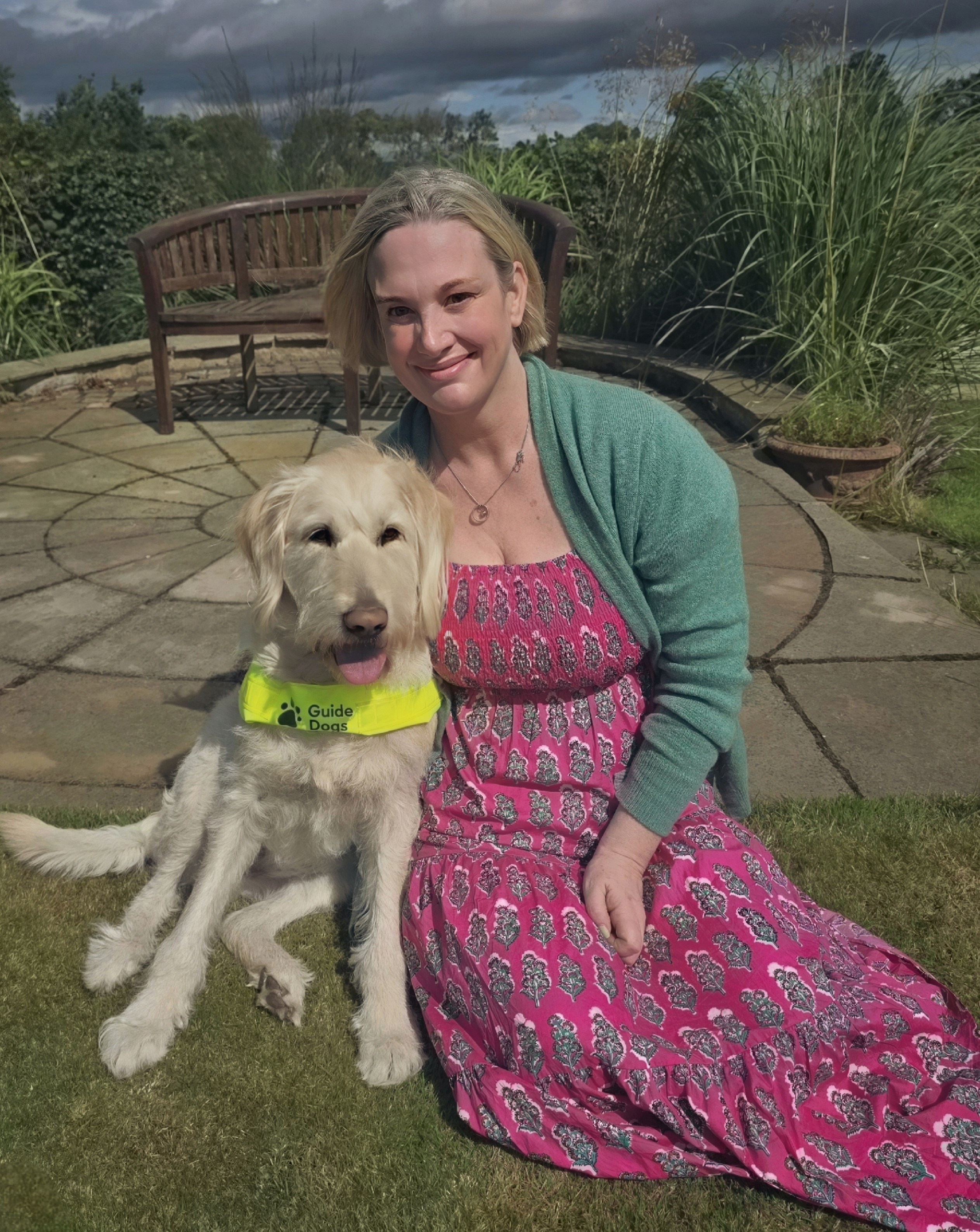 Helen wears a pink dress and green cardigan. She's sitting outside on the grass with her right arm around her Standard Poodle x Labrador Sunny wearing his guide dog harness.
