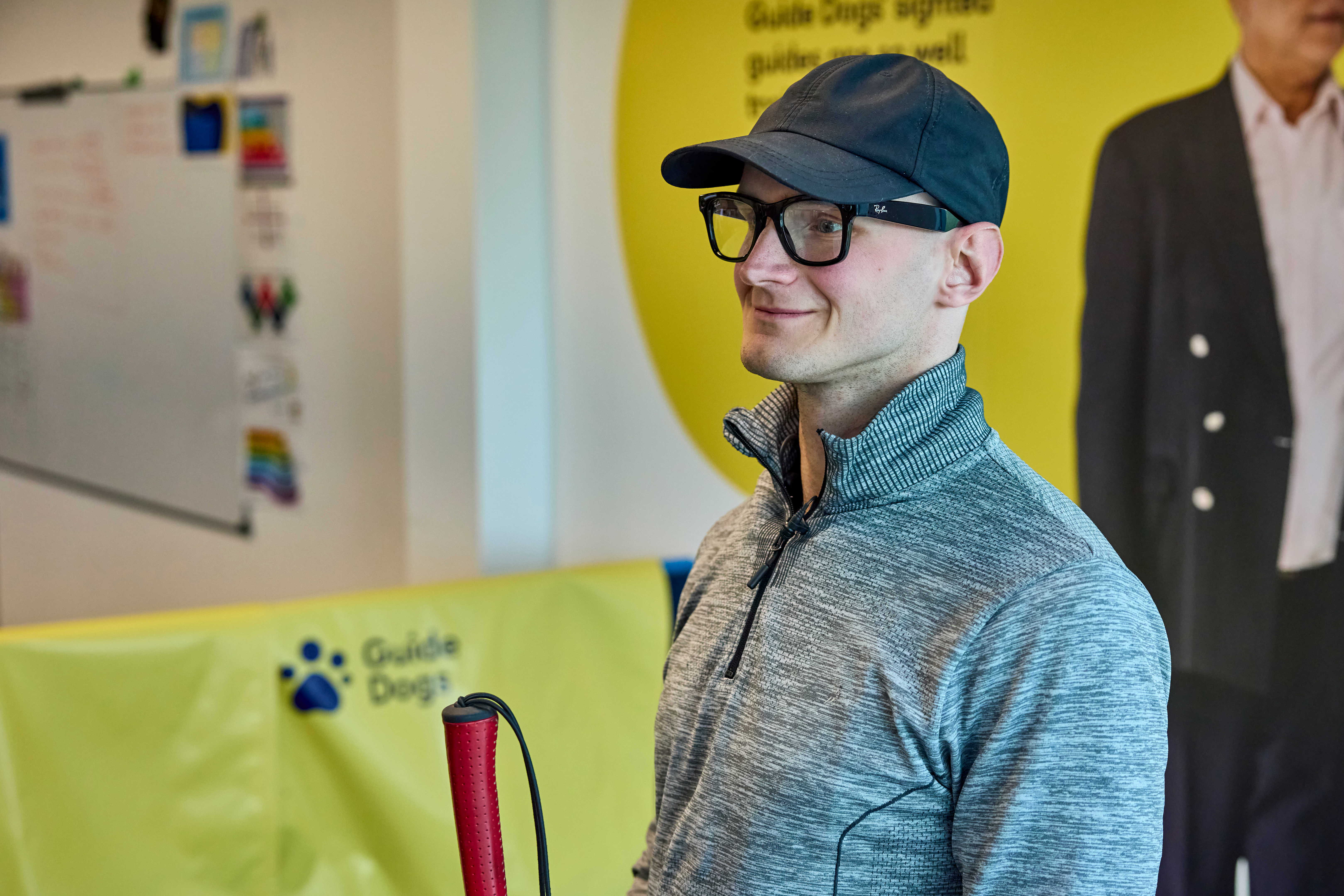 Peter, an assistive tech user, smiles. He's wearing a cap and a pair of Ray-Ban Meta glasses and holds his cane in one hand.